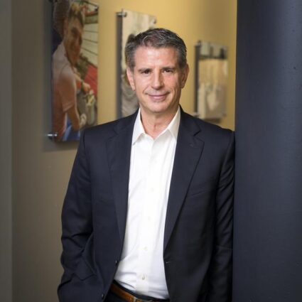 Marty is in a black suit jacket with a white shirt, leaning on a dark blue wall. In the background is a hallway with photos hanging up on the yellow wall.