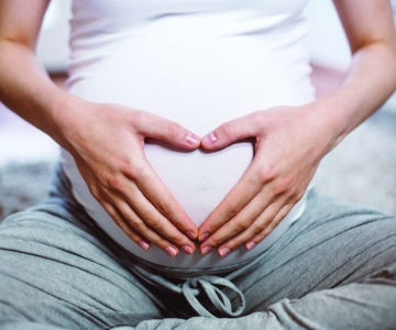 The belly of a pregnant person with their hands in the shape of a heart on their belly.