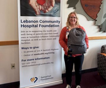 Alix Slayter stands indoors holding a gray food backpack in front of a banner for the Lebanon Community Hospital Foundation, which includes contact information and details about supporting patients.