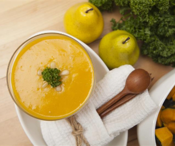 A bowl of creamy orange soup, garnished with parsley and a small cluster of white beans, sits on a light wood surface. Nearby are two yellow pears, a bundle of fresh kale, and a napkin wrapped around wooden serving spoons. A dish of roasted butternut squash pieces is partially visible on the side.