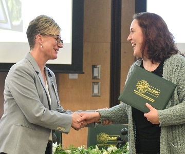 A short-haired woman wearing glasses in s sports coat shake the hand of a smiling lady with longer curly hair wearing a green sweater and holding an award.