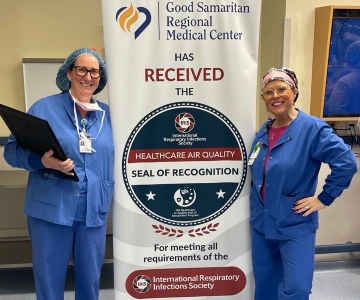 Two women stand next the recognition seal , Both are wearing glue scrubs one is holding an award in her hand.