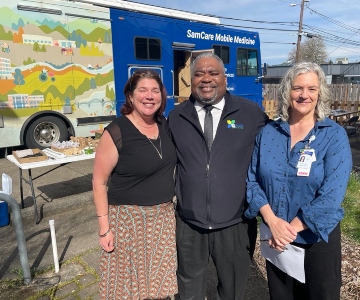 A man wearing a black jacket and tie stands in between two women one wearing a blue long sleeve shirt and the other wearing a black short sleeve blouse.