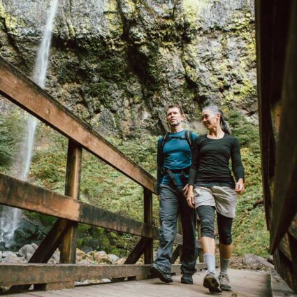 Man and woman hiking across a bridge with a waterfall in the background.
