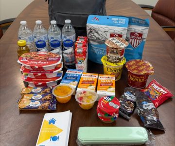A table displays the contents of a food backpack, including bottled water, freeze-dried meals, canned pasta, cereal cups, snack bars, fruit cups, pudding and informational pamphlets. A gray backpack is visible in the background.