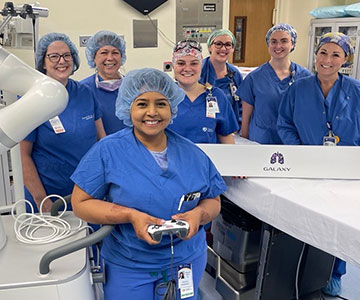 A woman dressed in blue scrubs holds a controller while six women stand behind her in an operating room.