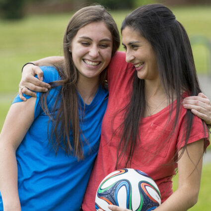 Smiling girls arms around shoulders with soccer ball.