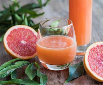 A clear glass filled with fresh pink grapefruit juice sits on a wooden surface, with a taller glass of juice partially visible behind it. Two halved grapefruits with vibrant pink flesh are placed nearby, along with green leaves scattered around for a natural, fresh look.