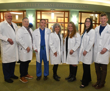 Seven clinicians wearing white coats standing in the lobby with green walls in the background.