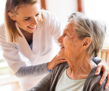 A smiling nurse leaning down to talk to an elderly patient with her hands on her patient's shoulders.