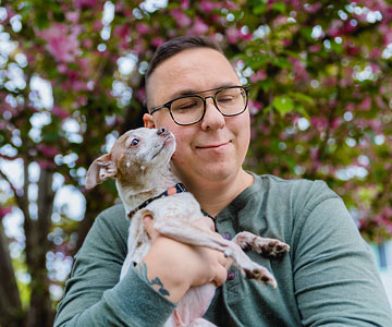 A smiling man standing underneath a flowering tree and hugging a small dog in his arms.