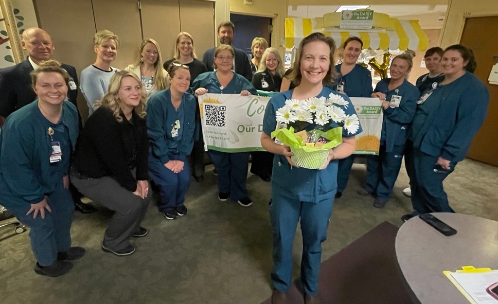 A woman holds a bouquet of daisies in front of a large group of men and women holding a sign.