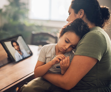 A mom with a sick child on her lap talks to a health care provider on a telehealth call through a tablet.