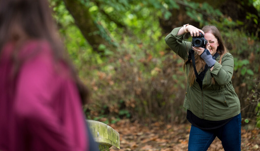Ashley smiling as she looks through her camera's viewfinder while taking a picture of her subject.
