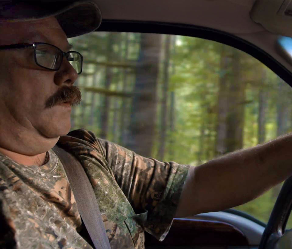 A close up of John behind the wheel of his truck as a forest of trees pass the window behind him.