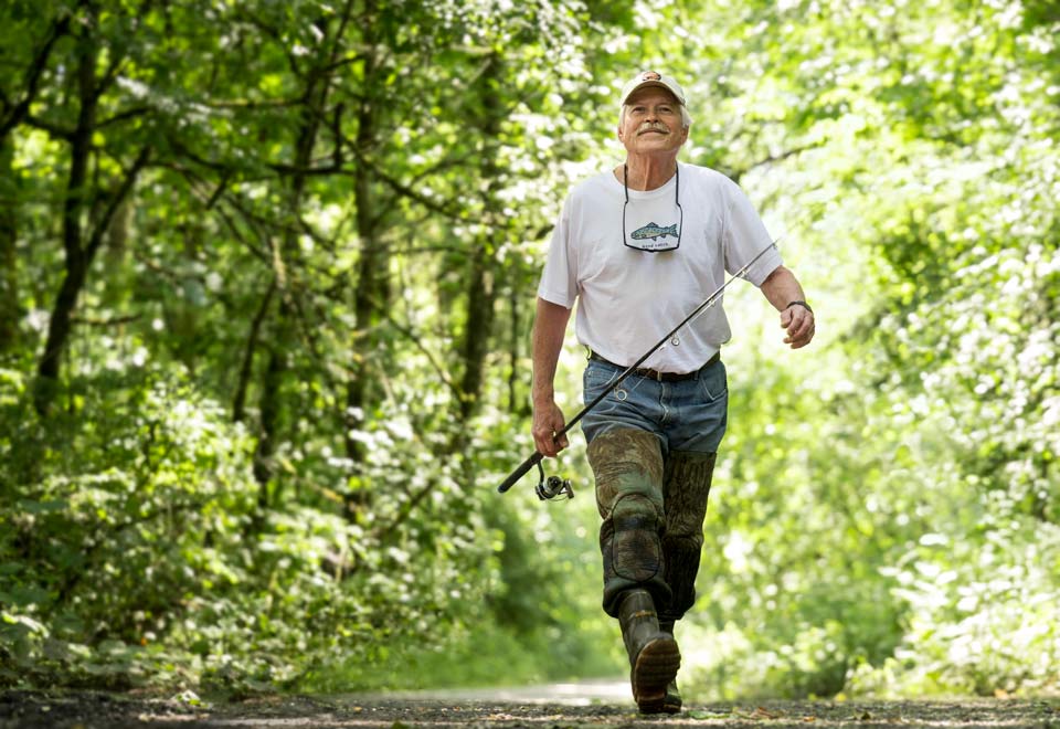 A smiling Randy walking down a path with his fishing pole in hand.