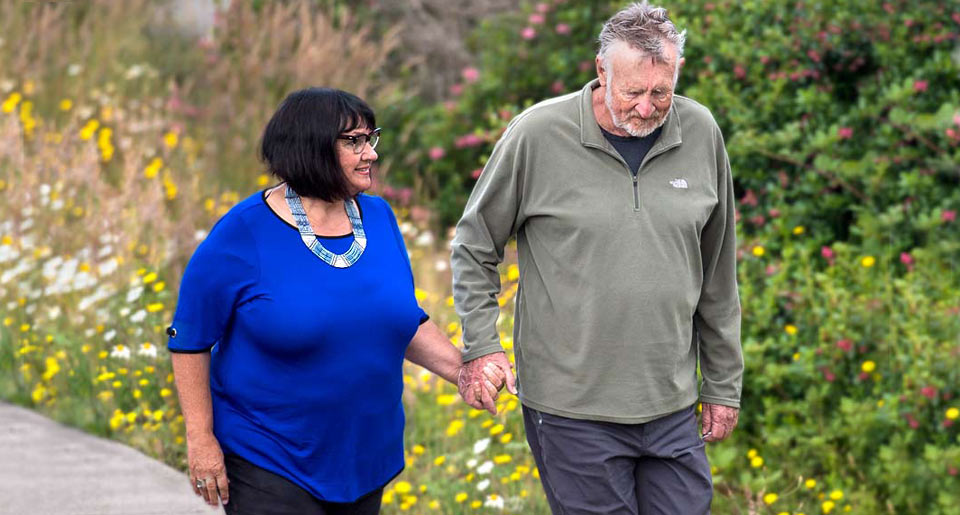 Vallie and Lynn walking hand in hand with bright yellow flowers blooming behind them.