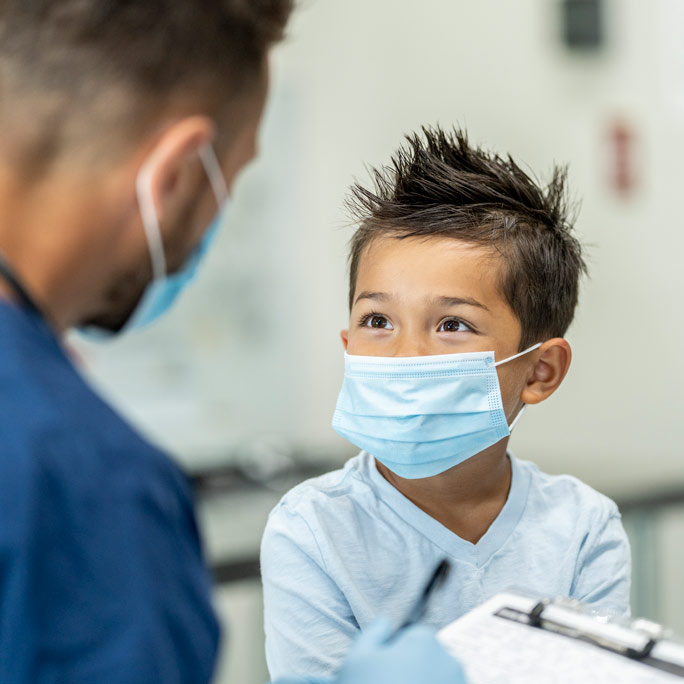 A young boy in a mask looking up at his health care provider.