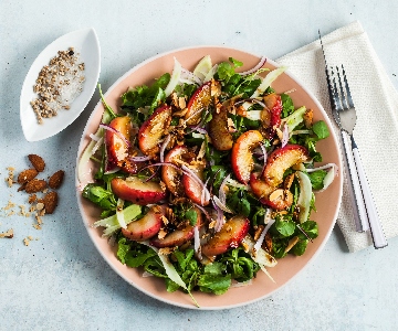 A fresh salad is served in a light pink bowl on a light gray surface. The salad includes mixed greens, sliced red onions, peach slices, and chopped nuts. To the left of the bowl is a small white dish with coarse salt and pepper, along with scattered almonds and seeds. On the right, a folded beige napkin holds a fork and knife.