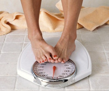 Close up of a woman's feet on a scale. Her toes of one foot rest over the other foot in a way that conveys self-consciousness.