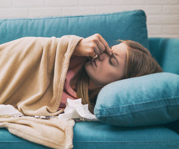 Woman laying on the cough pinching her nose and looking sick.