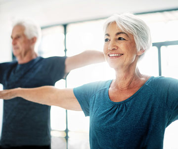 A smiling senior woman and man doing stretching exercise in a gym setting.