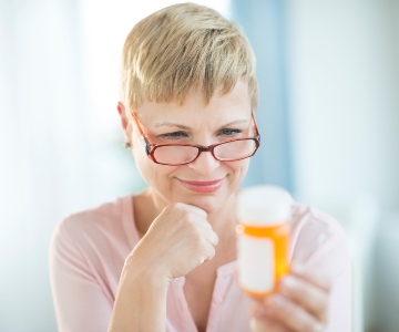 An older woman wearing glasses holds an orange pill bottle and reads the lable.