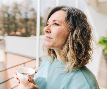 A woman looks out her window as she sips a cup of coffee.