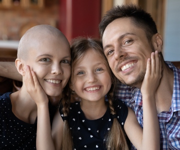 A young child smiling with her head between her parents. Mom is fighting cancer.